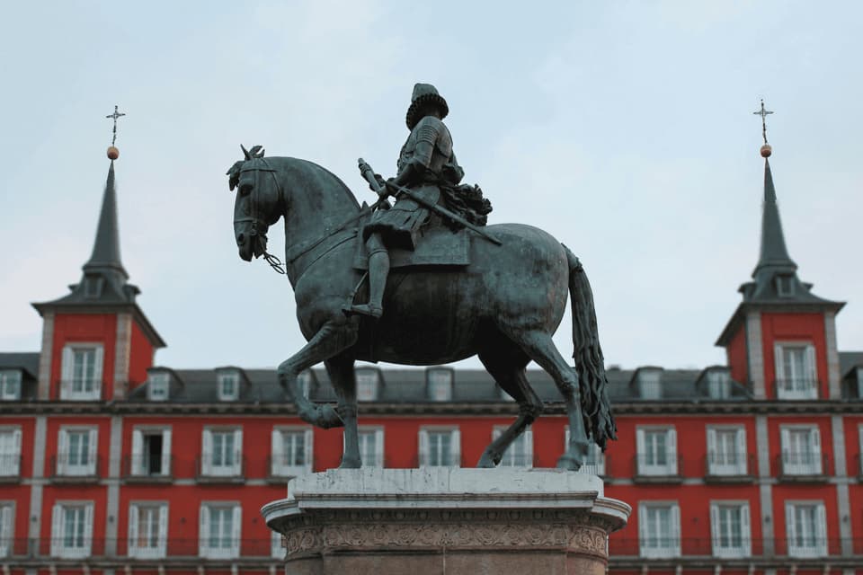 Une statue équestre en bronze sur un piédestal, avec un grand bâtiment rouge et ses tours jumelles en arrière-plan sous un ciel couvert.