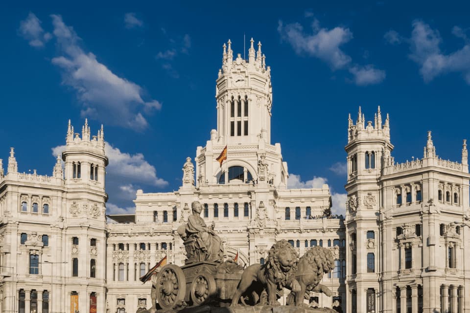 Une fontaine de pierre représentant un char tiré par des lions se dresse devant un palais blanc orné sous un ciel bleu nuageux.