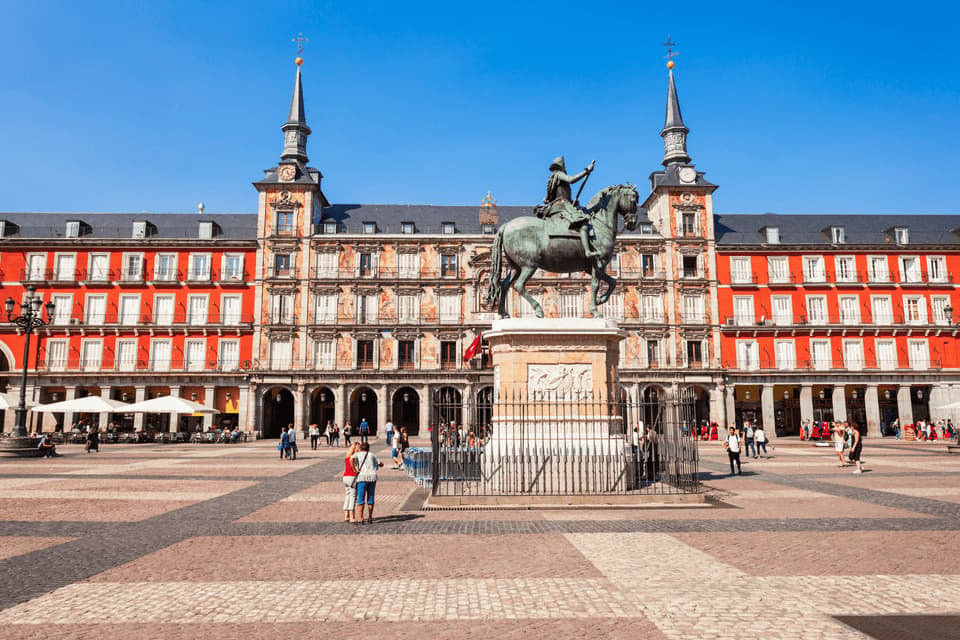 Une statue équestre en bronze trône sur un piédestal au centre d'une vaste place pavée, avec un bâtiment rouge historique en arrière-plan.