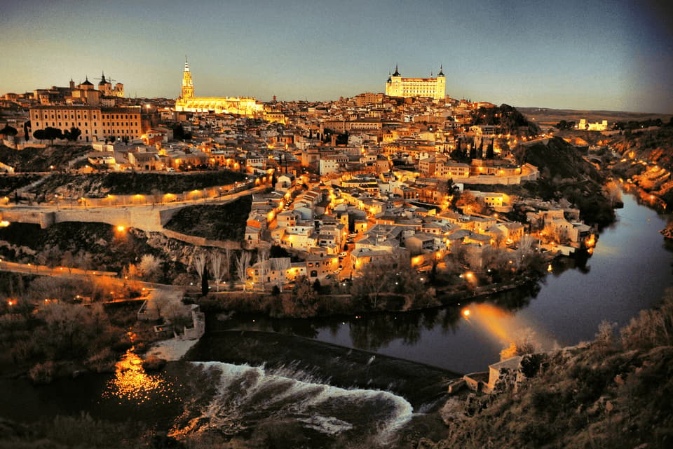 Vue panoramique d'une ville historique perchée, illuminée au crépuscule, avec une rivière et un petit déversoir au premier plan.