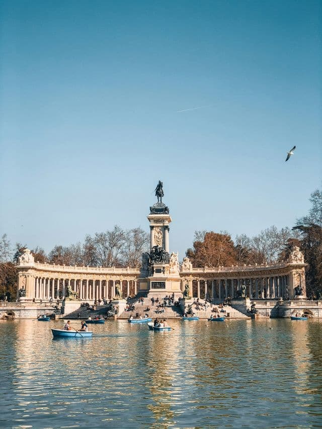 Des gens dans de petits bateaux bleus rament sur un lac devant un grand monument surmonté d'une statue équestre.