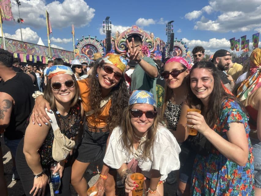 Un groupe d'amis WeRoad souriant pour un selfie à un festival de musique en plein air avec une scène colorée en arrière-plan.