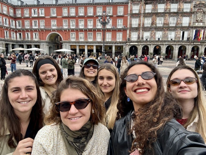Un voyage de groupe WeRoad de femmes souriantes prenant un selfie sur une place européenne animée avec des bâtiments historiques.