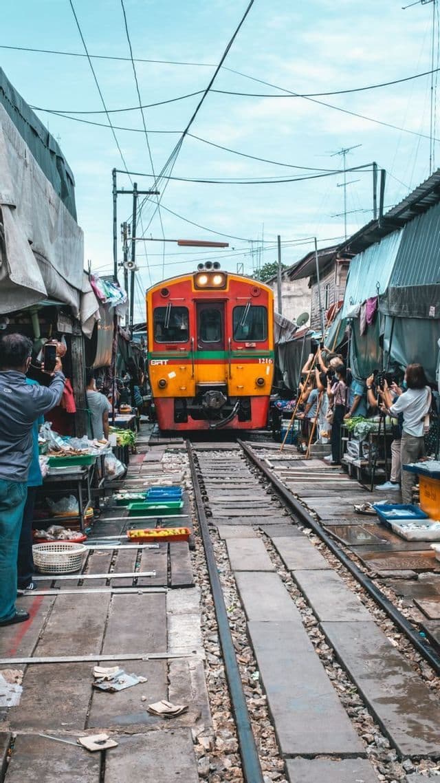 Un train orange et rouge s'approche sur les voies qui traversent un marché bondé, tandis que les gens s'écartent pour prendre des photos.