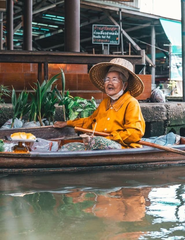 Una anciana vendedora con una camisa amarilla y un gran sombrero de paja se sienta en un bote de madera lleno de mercancías sobre el agua.