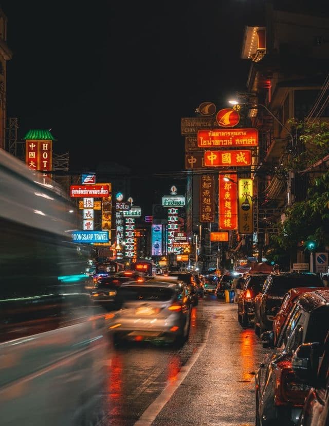 Une rue de ville animée la nuit, remplie de circulation automobile, illuminée par des néons colorés se reflétant sur le pavé mouillé.