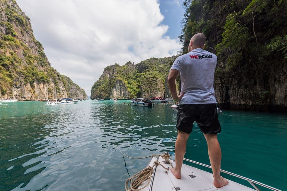 Un homme en t-shirt WeRoad se tient à la proue d'un bateau, regardant une lagune turquoise entourée de falaises rocheuses.