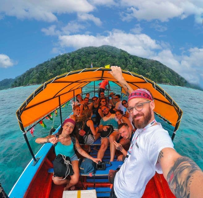 Ein Mann macht ein Selfie während einer WeRoad-Gruppenreise auf einem Boot mit orangem Verdeck, das auf türkisfarbenem Wasser nahe einer üppigen Insel segelt.
