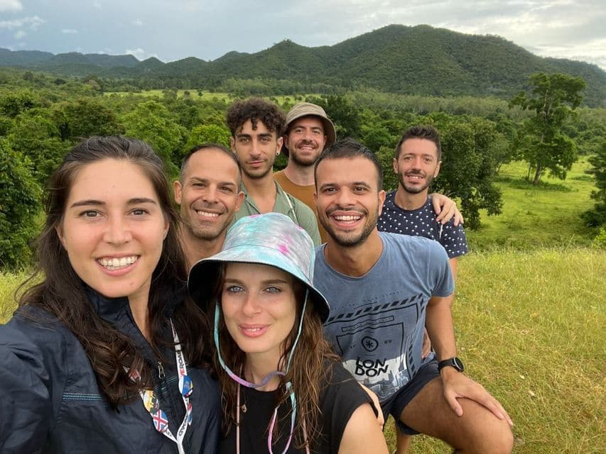 Un viaggio di gruppo WeRoad di sette persone sorridenti per un selfie in un campo erboso, con uno sfondo di verdi colline lussureggianti e alberi.