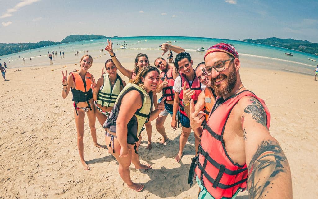Eine WeRoad-Gruppe macht mit Schwimmwesten ein lächelndes Selfie an einem sonnigen Sandstrand am Meer.