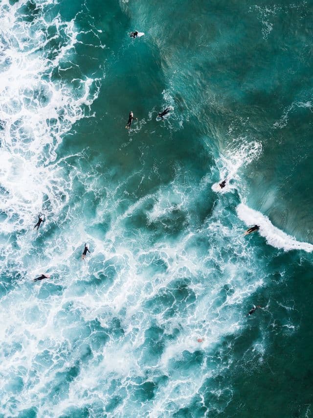 An aerial, top-down view of several surfers floating on their boards in turquoise ocean water among white, foamy waves.