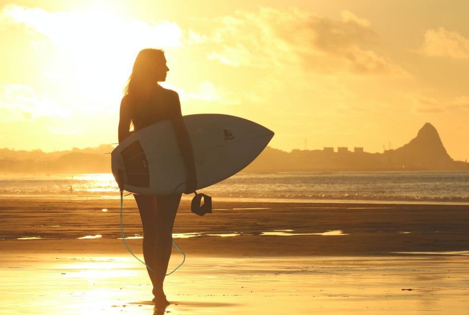A person in silhouette holds a surfboard while standing on a wet beach at sunset, with the ocean and a distant shoreline in view.