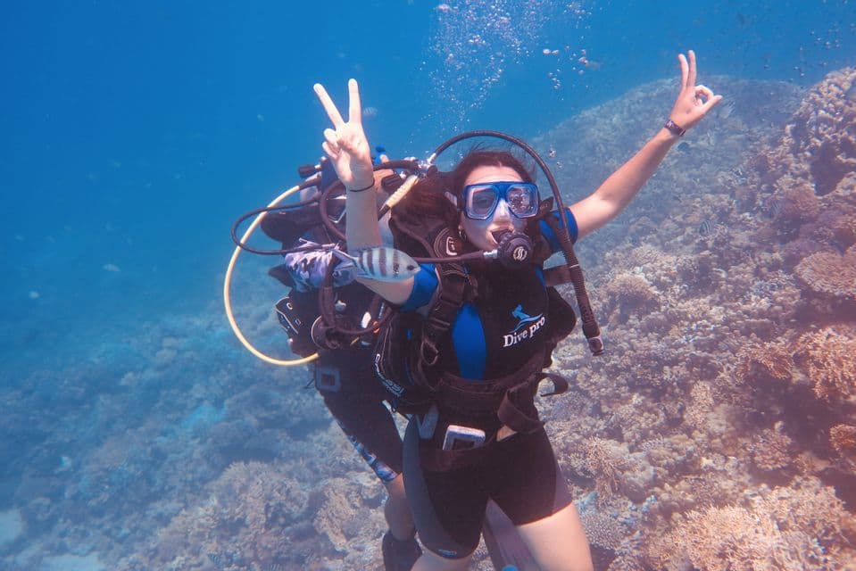 Una donna in attrezzatura da sub durante un viaggio di gruppo WeRoad fa i segni di pace e ok mentre si immerge vicino a una barriera corallina.
