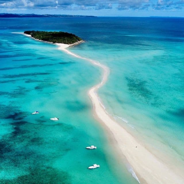 Una vista aerea di un banco di sabbia bianco serpeggiante che si estende da un'isola boscosa nell'oceano turchese, con diverse barche ancorate nelle vicinanze.