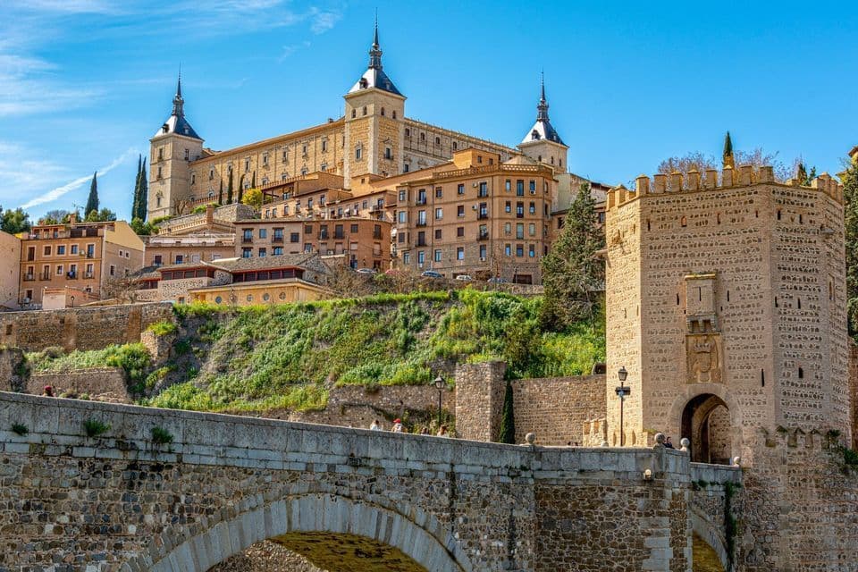 Un grande castello di pietra sorge su una verde collina, sovrastando un ponte di pietra ad arco con una torre d'ingresso, sotto un cielo blu e soleggiato.