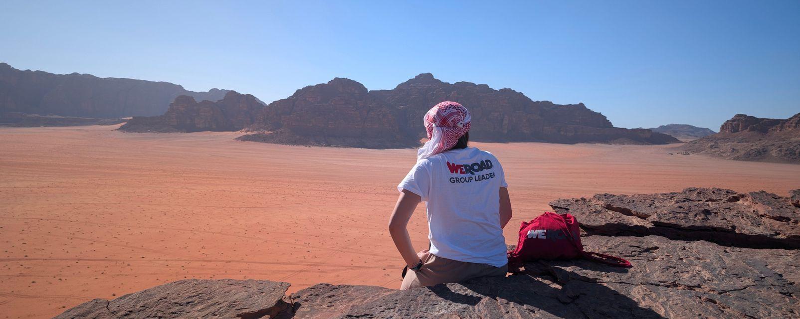Un capogruppo WeRoad con una kefiah è seduto su una sporgenza rocciosa, ammirando un vasto paesaggio desertico rosso e montagne lontane.