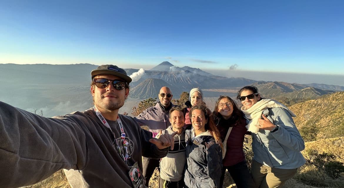 Un viaggio di gruppo WeRoad scatta un selfie su un punto panoramico di montagna con vulcani attivi che fumano sullo sfondo sotto un cielo sereno.