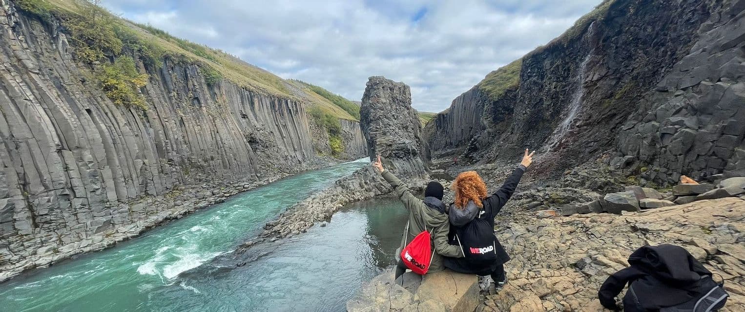 Da dietro, due persone di un viaggio di gruppo WeRoad sono sedute su una roccia che si affaccia su un fiume che scorre in un canyon di colonne basaltiche.