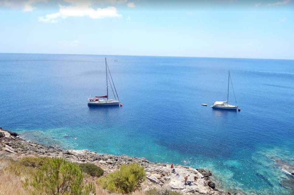 Due barche a vela ancorate in acque calme e blu vicino a una costa rocciosa dove un gruppo WeRoad nuota e si rilassa.