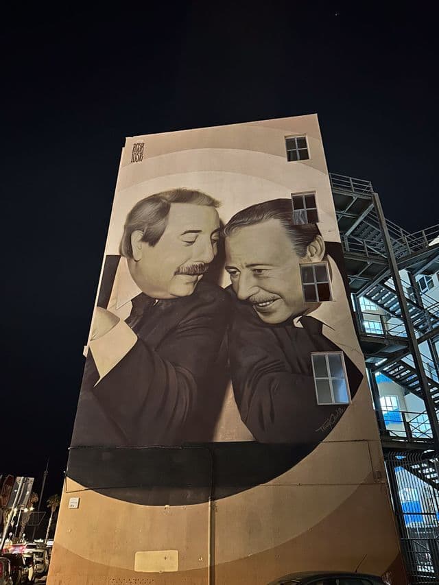 Un grande murale in bianco e nero di due uomini sorridenti con i baffi dipinto sul lato di un edificio di notte.