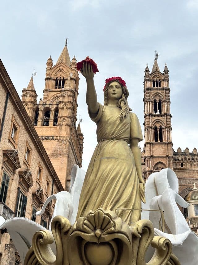 Una statua dorata di una donna con una corona di fiori regge un fiore rosso davanti a torri cattedrali ornate sotto un cielo nuvoloso.