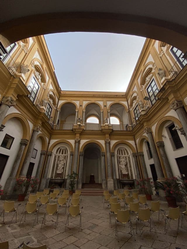 Un'ampia inquadratura dal basso di un cortile a cielo aperto di un sontuoso edificio, con file di sedie gialle sul pavimento in pietra.