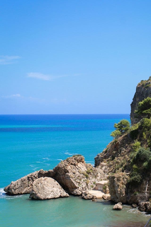 Una costa rocciosa incontra un mare turchese sotto un cielo azzurro limpido, con scalini in pietra che conducono a una piccola caletta appartata.