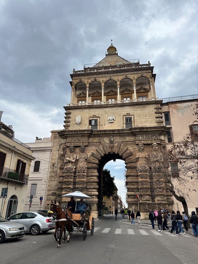 Una carrozza trainata da cavalli viaggia su una strada davanti a un grande arco in pietra ornato sotto un cielo nuvoloso.