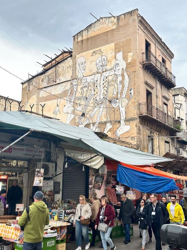 Un gruppo WeRoad attraversa un vivace mercato all'aperto, con un grande murale artistico dipinto sulla facciata di un vecchio edificio.