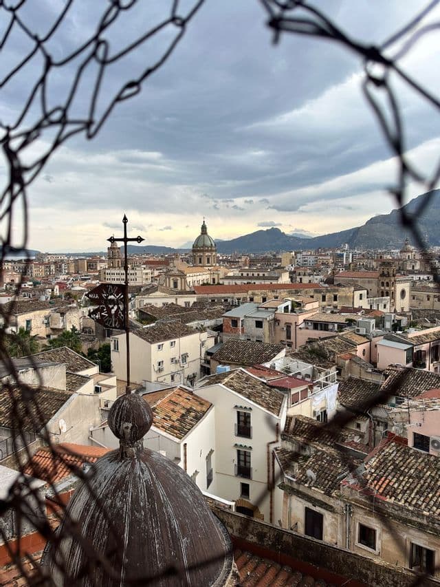 Veduta dall'alto di una città con tetti in terracotta e una grande cupola, intravista attraverso un foro in una recinzione metallica sotto un cielo nuvoloso.