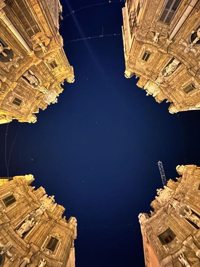 Una prospettiva dal basso, che inquadra quattro angoli di edifici illuminati e riccamente decorati, incorniciando un cielo notturno buio e stellato.