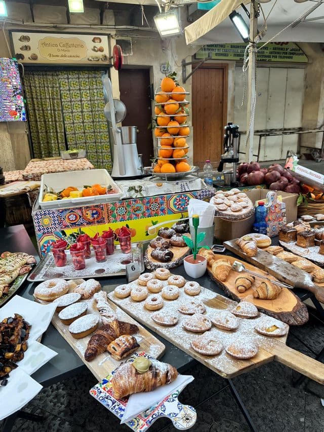Una bancarella di cibo all'aperto con un'ampia esposizione di pasticcini, dolci, arance fresche e coppette di frutta sui tavoli.