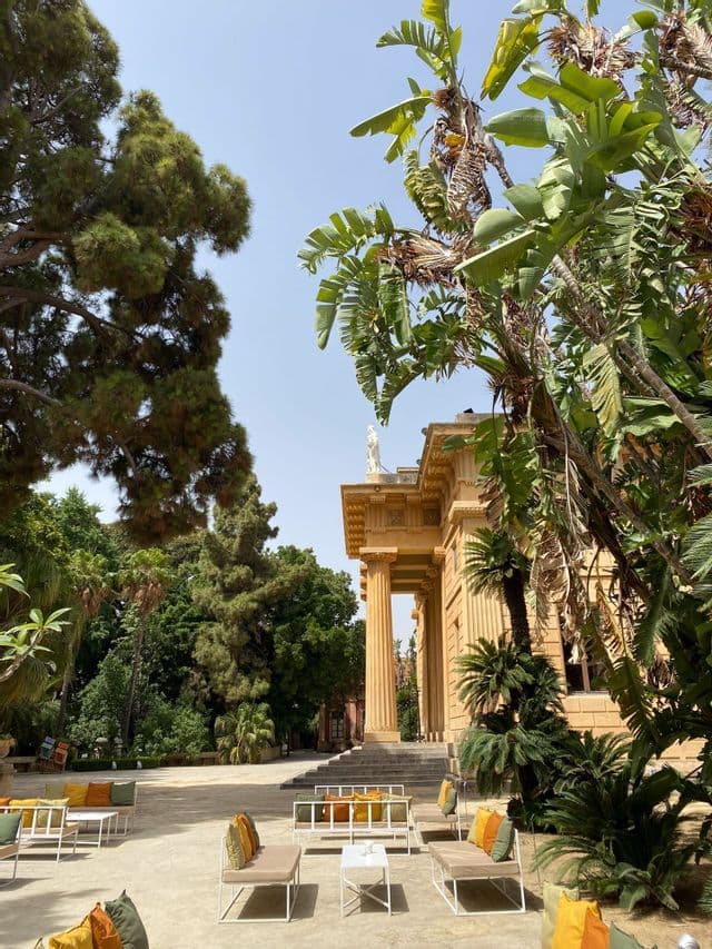 Cortile soleggiato con posti a sedere moderni all'aperto, di fronte a un edificio classico con colonne, circondato da alberi lussureggianti.