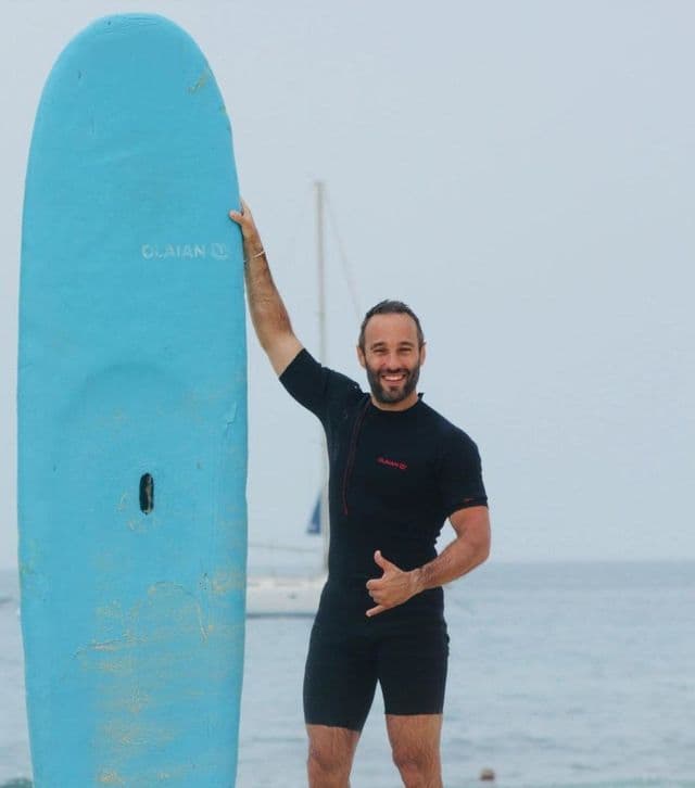 Un hombre sonriente con traje de neopreno negro se encuentra en una playa, sosteniendo una tabla de surf azul y haciendo el gesto shaka.