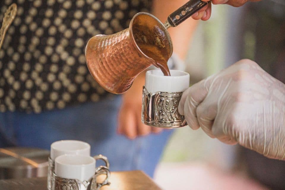 Una persona vierte café caliente de un cezve de cobre tradicional en una pequeña taza blanca sostenida por una mano enguantada.