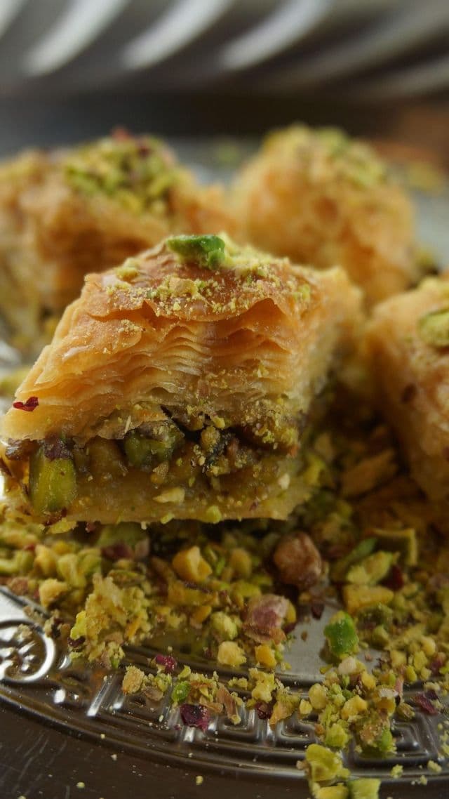 Un primer plano de un trozo de baklava mostrando capas de hojaldre y un relleno de pistacho, cubierto con frutos secos triturados en un plato.