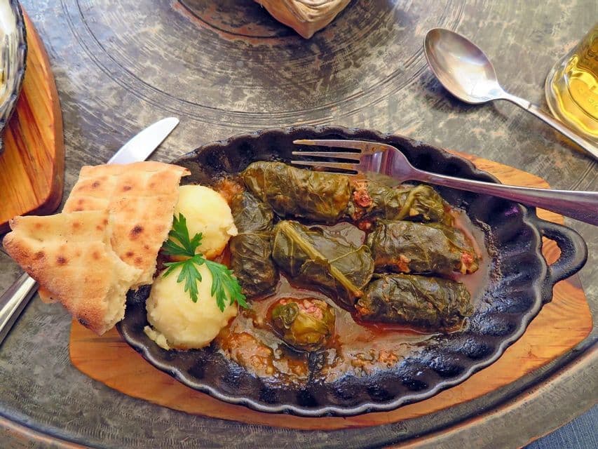 Un plato de hojas de parra rellenas en una fuente negra, acompañado de puré de patatas, pan plano y un tenedor en una bandeja metálica.