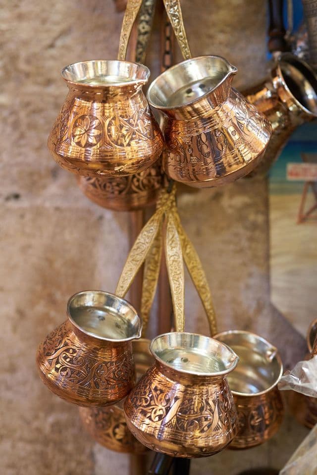 Primer plano de varias cafeteras turcas de cobre, grabadas y ornamentadas, colgadas para la venta en una pared de piedra.