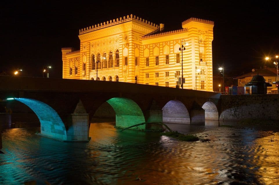 Un edificio ornamentado, iluminado en amarillo, se alza tras un puente de piedra sobre un río por la noche, con sus arcos iluminados en azul y verde.
