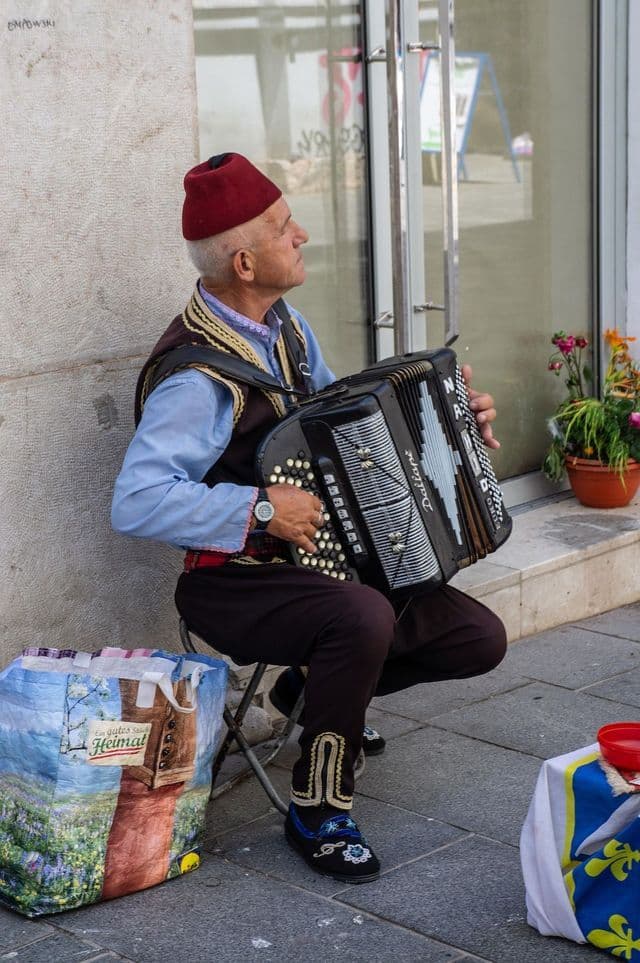 Un anciano con ropa tradicional, incluyendo un fez rojo y un chaleco bordado, está sentado en un taburete en una acera tocando el acordeón.