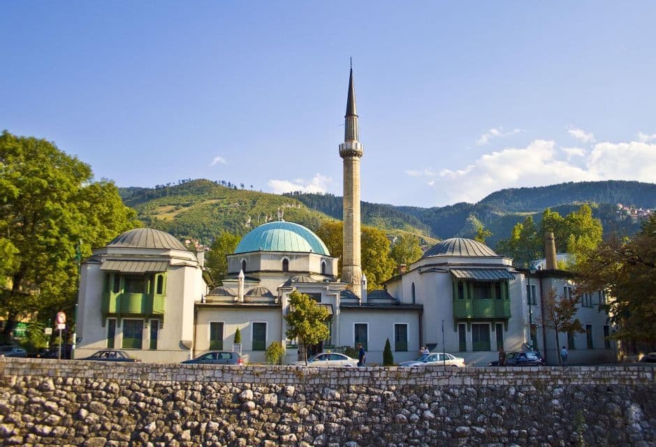 Una mezquita con cúpula turquesa y un alto minarete se alza frente a exuberantes colinas verdes bajo un cielo azul brillante.