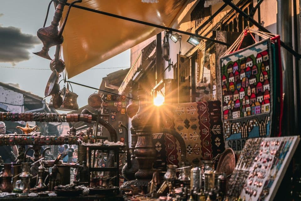 Un puesto de mercado al aire libre exhibe ollas de cobre y alfombras coloridas bajo un toldo, con un brillante destello de sol iluminando el callejón.