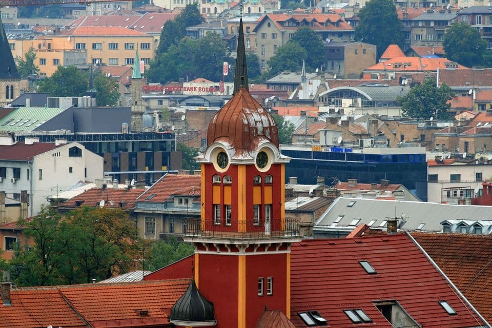 Una torre roja y amarilla con una cúpula de cobre y una aguja puntiaguda se eleva sobre un denso paisaje urbano con tejados de tejas rojas.