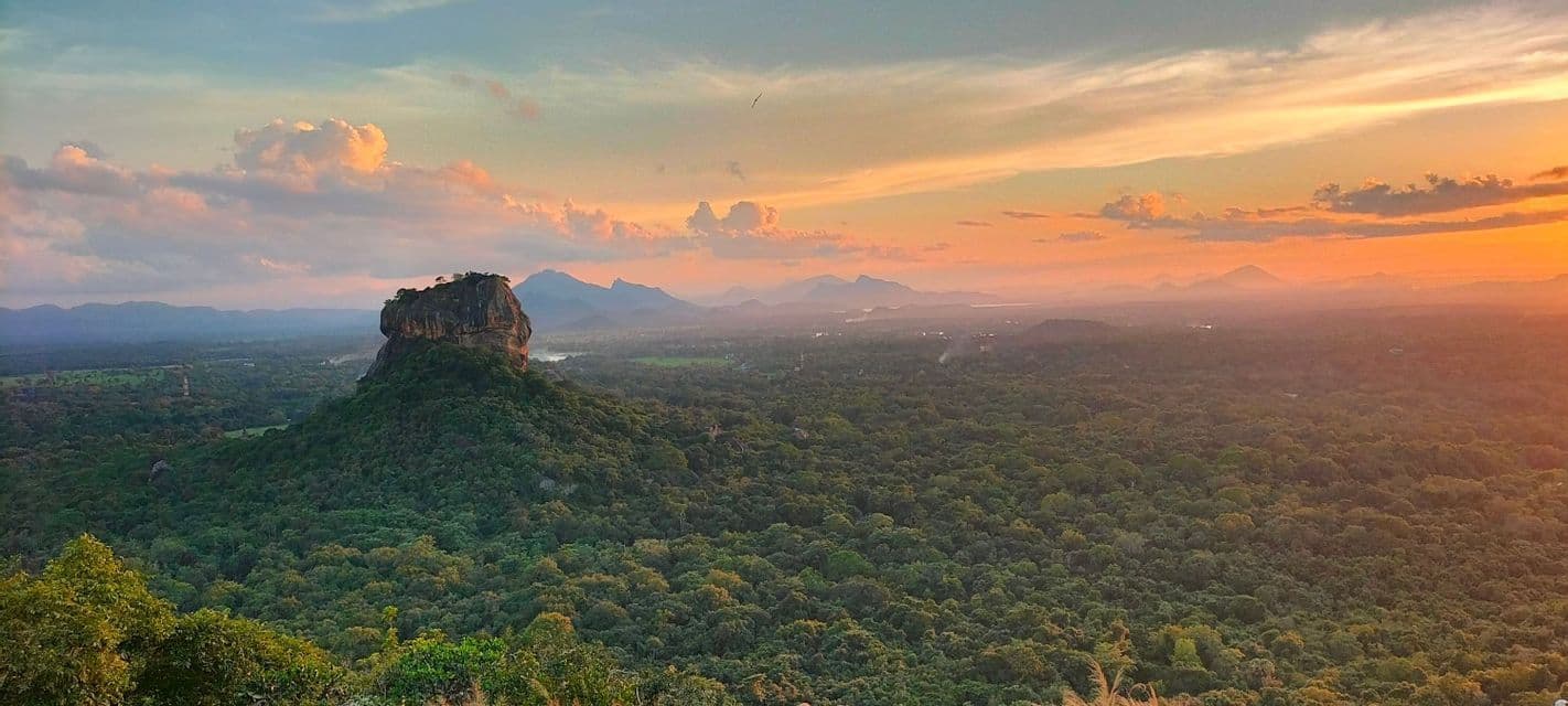 Panoramica di un'imponente formazione rocciosa che emerge da una vasta foresta verde, sotto un cielo colorato al tramonto, con montagne in lontananza.