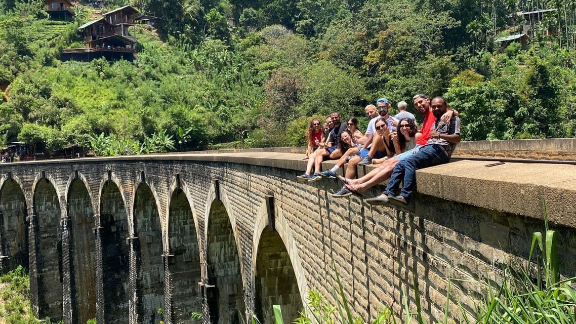Un gruppo WeRoad in viaggio, seduto insieme sul bordo di un grande ponte ad arco in pietra, in una lussureggiante valle verde.