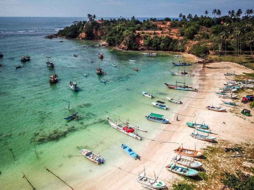Vista aerea di numerose barche da pesca ormeggiate in una baia tropicale con acqua turchese cristallina e una spiaggia di sabbia.