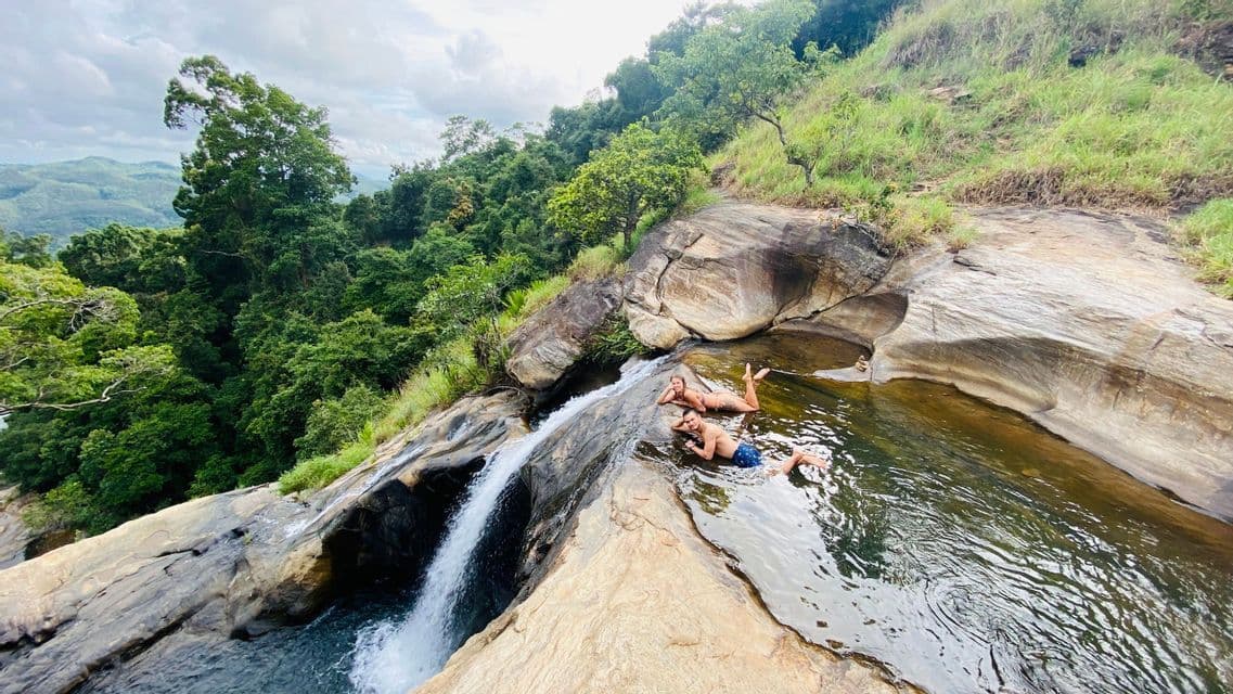 Un uomo e una donna si rilassano in una piscina naturale rocciosa poco profonda ai margini di una cascata, con vista su una fitta foresta verde.