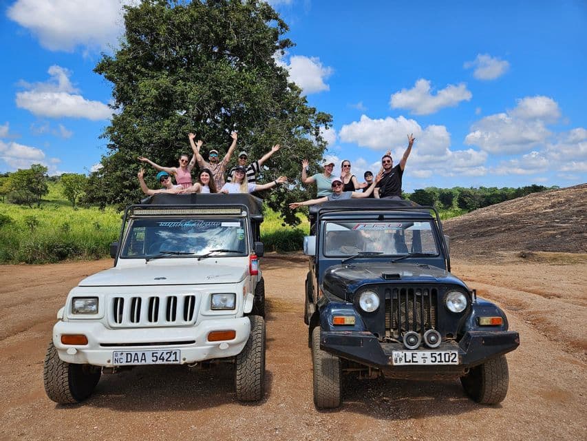 Un viaggio di gruppo WeRoad sorride e saluta dai tetti aperti di due jeep da safari parcheggiate su una strada sterrata sotto un cielo azzurro.