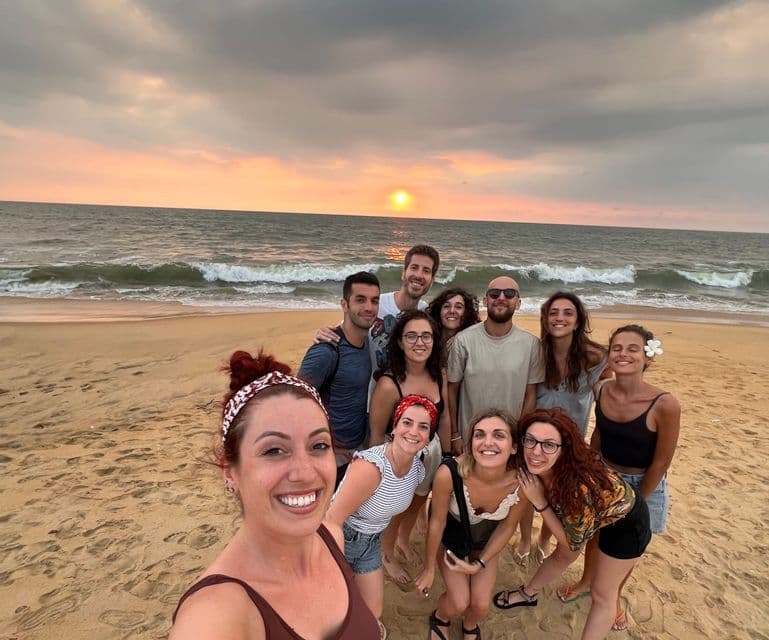 Un viaggio di gruppo WeRoad si fa un selfie su una spiaggia sabbiosa mentre il sole tramonta sull'oceano.