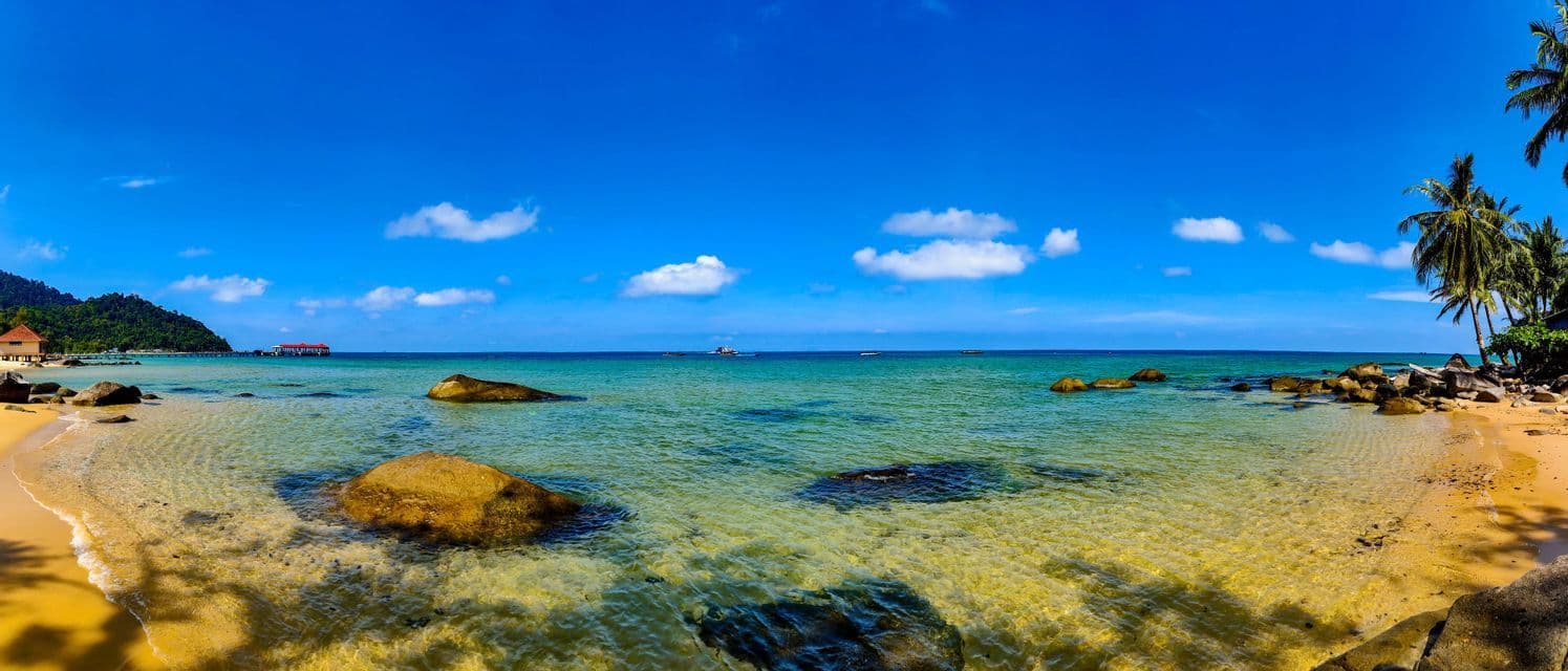 Vista panoramica di una spiaggia tropicale con acqua turchese cristallina, grandi rocce sulla sabbia e un pontile lontano sotto un cielo azzurro intenso.